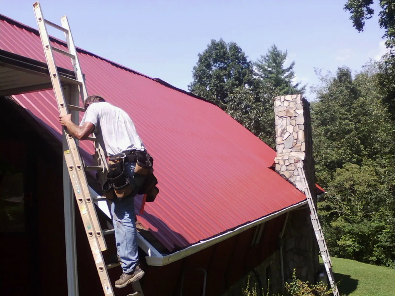 Roofer on a ladder beside a red metal roof and stone chimney — Soffit & Fascia Repair by Roofing Brothers in Haledon